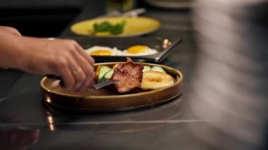 Profesyonel bir mutfakta, temiz metal bir masanın üstünde kadın bir şef yuvarlak ahşap bir tabakta İngiliz kahvaltısı toplar.