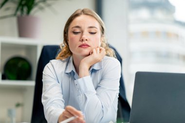 Modern bir ofiste büyük bir penceresi olan mavi gömlekli, dizüstü bilgisayarlı güzel bir bayan işçi masada oturur sıkıcı bir iş başını dik tutar ve günün sonunu bekler.