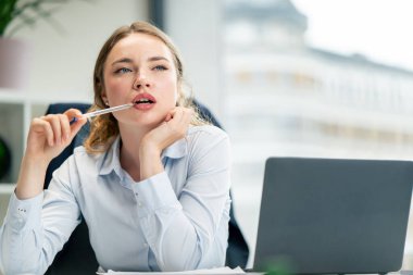 Modern bir ofiste, geniş pencereli, mavi gömlekli, dizüstü bilgisayarlı güzel bir işçi elinde kalemle hayallere dalgın dalgın dalgın oturuyor.