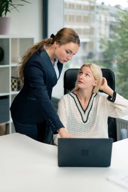 Geniş pencereli modern bir ofiste mavi gömlekli güzel bir işçi kıdemli bir uzman, genç bir uzmana dizüstü bilgisayar öğrencisiyle nasıl çalışacağını öğretiyor.