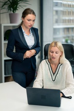 Geniş pencereli modern bir ofiste mavi gömlekli güzel bir işçi kıdemli bir uzman, genç bir uzmana dizüstü bilgisayar öğrencisiyle nasıl çalışacağını öğretiyor.
