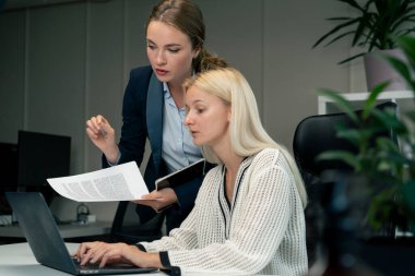 Geniş pencereli modern bir ofiste mavi gömlekli güzel bir işçi kıdemli bir uzman, genç bir uzmana dizüstü bilgisayar öğrencisiyle nasıl çalışacağını öğretiyor.