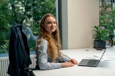 Geniş pencereli modern bir ofiste mavi gömlekli güzel bir işçi kameraya bakar, genç bir patron.