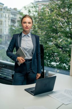 Modern bir ofiste, geniş pencereli güzel bir bayan işçi mavi gömlekli, mavi takım elbiseli, elinde laptop ve tabletle kameraya bakıyor.