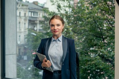 Modern bir ofiste, geniş pencereli güzel bir bayan işçi mavi gömlekli, mavi takım elbiseli, elinde laptop ve tabletle kameraya bakıyor.
