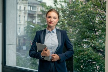 Modern bir ofiste, geniş pencereli güzel bir bayan işçi mavi gömlekli, mavi takım elbiseli, elinde laptop ve tabletle kameraya bakıyor.