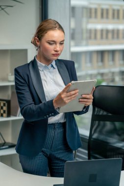 Modern bir ofiste, büyük bir penceresi olan güzel bir bayan işçi elinde tabletle mavi bir takım elbiseyle duruyor ve ayın çalışma planına bakıyor.
