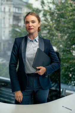 Modern bir ofiste büyük bir penceresi olan güzel bir bayan işçi elinde dizüstü bilgisayarla kameraya bakıyor.