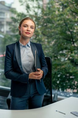 Modern bir ofiste büyük bir penceresi olan güzel bir bayan işçi elinde dizüstü bilgisayarla kameraya bakıyor.