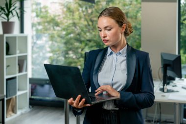 Modern bir ofiste büyük bir penceresi olan güzel bir bayan işçi elinde dizüstü bilgisayarla mavi bir takım elbise içinde ayın çalışma planına bakıyor.