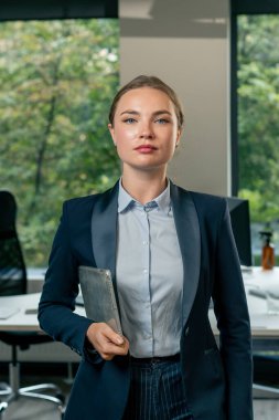 Modern ofisin ortasında büyük bir pencereyle mavi gömlekli ve elinde tabletli mavi takım elbisesi olan güzel bir bayan işçi kameraya bakıyor.