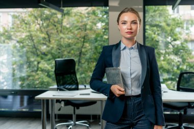 Modern ofisin ortasında büyük bir pencereyle mavi gömlekli ve elinde tabletli mavi takım elbisesi olan güzel bir bayan işçi kameraya bakıyor.