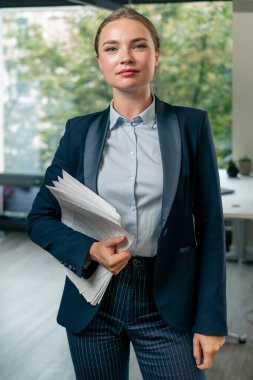 Merkezde geniş pencereli, modern bir ofiste mavi takım elbiseli güzel bir işçi elinde belgelerle genç bir ekip bekliyor.