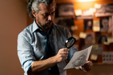 Dedektiflik bürosunda gömlekli ve kravatlı orta yaşlı bir dedektif büyüteçle inceler kanıt inceler suçlu fotofit güzergahı suçlu tahtası ipuçlarını inceler