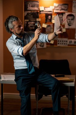 Dedektiflik bürosunda gömlekli ve kravatlı orta yaşlı bir dedektif büyüteçle inceler kanıt inceler suçlu fotofit güzergahı suçlu tahtası ipuçlarını inceler
