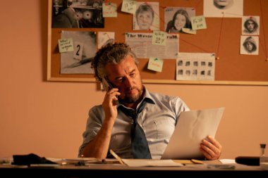 Dedektiflik bürosuna yakın plan. Gömlek ve kravat takmış orta yaşlı bir dedektif. Masada oturmuş fotofite bakıyor ve telefonla konuşuyor.