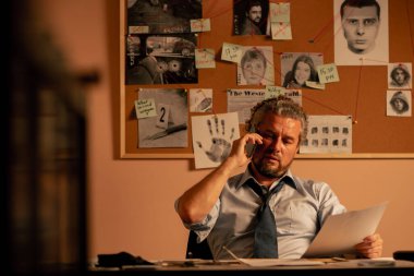 Dedektiflik bürosuna yakın plan. Gömlek ve kravat takmış orta yaşlı bir dedektif. Masada oturmuş fotofite bakıyor ve telefonla konuşuyor.