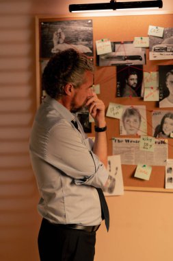 Dedektif ofisini kapatın. Gömlekli ve kravatlı orta yaşlı araştırmacı gerçeği bulmak için ipucu panosunun yanında duruyor.