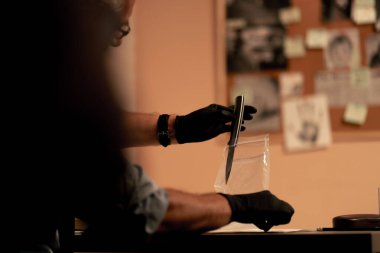 Yakın plan dedektiflik bürosu gömlekli ve kravatlı orta yaşlı dedektif masasında oturmuş kanlı bıçaklı cinayet silahını inceliyor.