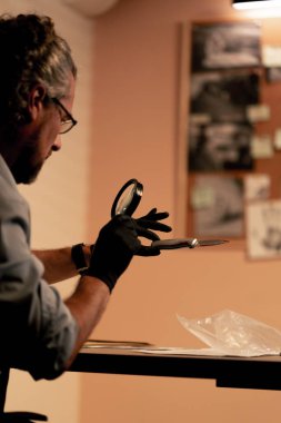 Yakın plan dedektiflik bürosu gömlekli ve kravatlı orta yaşlı dedektif masasında oturmuş kanlı bıçaklı cinayet silahını inceliyor.