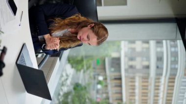 Modern bir ofiste, geniş pencereli dikey bir video. Mavi tişörtlü güzel bir işçi. Dizüstü bilgisayarlı bir konuşma.