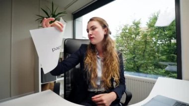 Modern bir ofiste geniş pencereli, mavi gömlekli güzel bir bayan çalışan kameraya doğrudan bakıyor ve beyaz bir sayfa yırtıyor.