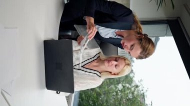 Modern bir ofiste geniş pencereli dikey bir video mavi gömlekli güzel bir işçi kıdemli bir uzman, genç bir uzmana dizüstü bilgisayar öğrencisiyle nasıl çalışacağını öğretiyor.