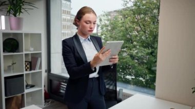 Modern bir ofiste, büyük bir penceresi olan güzel bir bayan işçi elinde tabletle mavi bir takım elbiseyle duruyor ve ayın çalışma planına bakıyor.