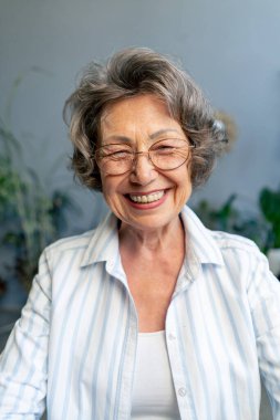 Çok yaşlı, gri saçlı, açık renk bluzlu bir büyükannenin yakın plan resmi. Hafif bir gülümsemesi var. Mavi duvarın arkasında oturuyor.