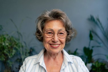 Çok yaşlı, gri saçlı, açık renk bluzlu bir büyükannenin yakın plan resmi. Hafif bir gülümsemesi var. Mavi duvarın arkasında oturuyor.