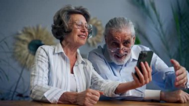 Yaşlı insanlar bir masada oturup sosyal ağları izliyorlar komik videolar, mutlu yaşlı çiftler, güzel anılar, yaşlılıktan sonra sıcak duygular destek oluyor.