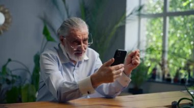 Çok yaşlı, gri saçlı, mavi tişörtlü, hafif gülüşlü, mavi bir duvara yaslanmış, telefonda mesajlaşan büyük ev bitkileriyle.