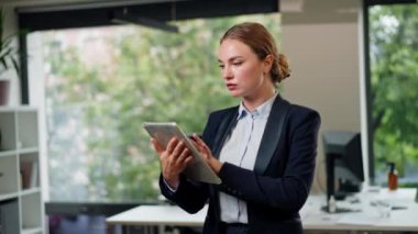 Modern bir ofiste, büyük bir penceresi olan güzel bir bayan işçi elinde tabletle mavi bir takım elbiseyle duruyor ve ayın çalışma planına bakıyor.