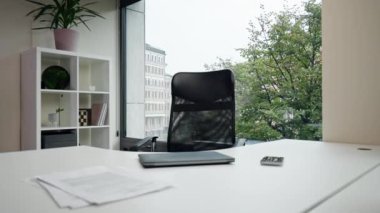 Modern bir ofiste, beyaz bir masanın yanında dizüstü bilgisayarı olan büyük bir pencere var. Siyah bir ofis koltuğu var. Rahat çalışma, sandalyenin arkası esnek.