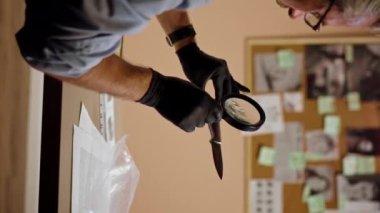 dikey video yakın plan dedektiflik bürosu gömlekli ve kravatlı orta yaşlı dedektif masasında oturmuş kanlı bıçaklı cinayet silahını inceliyor.