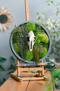 Close-up in a large green workshop against the background of a light wall there is a small easel with a round green decor made of stabilized moss and bird cherry of a small animal, hand-made robot clipart