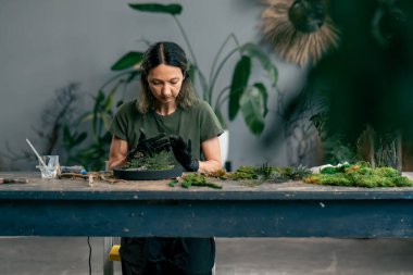 Büyük yeşil bir atölyede, yeşil saçlı, siyah eldivenli bir kadın büyük bir masada sabitlenmiş yosunlardan bir kompozisyon topluyor.