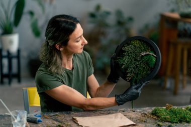 in a large green workshop, a thin woman with green hair in black gloves at a large table collects a composition from stabilized moss clipart