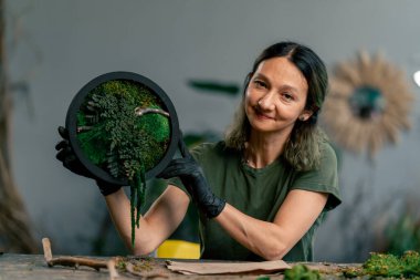 Büyük yeşil bir atölyede, ahşap bir masada yeşil saçlı zayıf bir kadın kameraya sabitlenmiş yosun bileşimini gösterir.