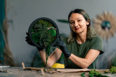 Büyük yeşil bir atölyede, ahşap bir masada yeşil saçlı zayıf bir kadın kameraya sabitlenmiş yosun bileşimini gösterir.