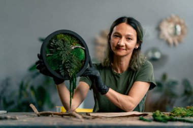 Büyük yeşil bir atölyede, ahşap bir masada yeşil saçlı zayıf bir kadın kameraya sabitlenmiş yosun bileşimini gösterir.