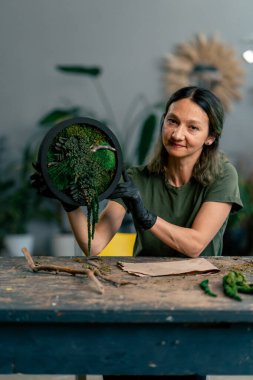 Büyük yeşil bir atölyede, ahşap bir masada yeşil saçlı zayıf bir kadın kameraya sabitlenmiş yosun bileşimini gösterir.