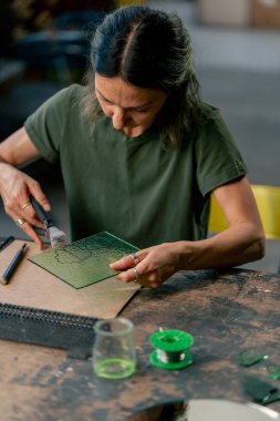 Büyük yeşil bir atölyede, yeşil saçlı ince bir kadın büyük bir masada yeşil yaprakları kesiyor, tasarımı çiziyor, şekillendiriyor.