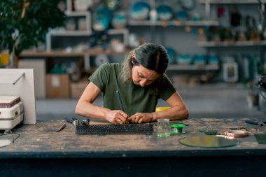 Büyük yeşil bir atölyede, yeşil saçlı ince bir kadın büyük bir masada yeşil yaprakları kesiyor, tasarımı çiziyor, şekillendiriyor.