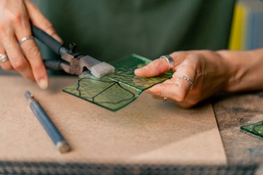 Büyük yeşil bir atölyeye yakın büyük bir masada yeşil saçlı zayıf bir kadın camdan yeşil taç yaprakları kesiyor ve bir tasarım çiziyor.