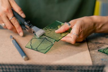 Büyük yeşil bir atölyeye yakın büyük bir masada yeşil saçlı zayıf bir kadın camdan yeşil taç yaprakları kesiyor ve bir tasarım çiziyor.