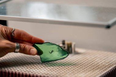 Büyük yeşil bir atölyeye yakın, öğütme makinesinin ahşap masasında yeşil saçlı, ince bir kadın cam bir ürünün uçlarını damak, köşeleri cilalıyor, el yapımı.