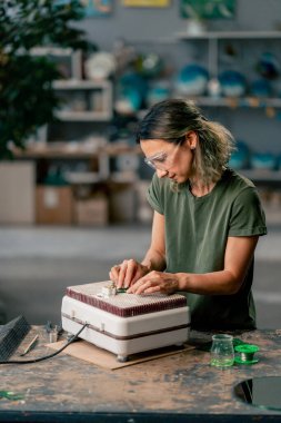 Büyük yeşil bir atölyede, öğütme makinesinin ahşap masasında yeşil saçlı, ince bir kadın cam bir ürünün ucunu, köşelerini cilalıyor, el yapımı.