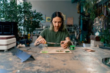 Yeşil saçlı bir kadın üzerinde büyük yeşil bir atölyede dekoratif çiçekler yaratıyor ayna el sanatlarında metal bant dekoru ile çalışıyor.