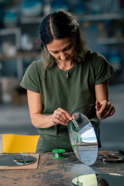 Büyük yeşil bir atölyede, yeşil saçlı bir kadın kameraya yansıyan cam çiçekle aynayı süslemeyi bitirdi.